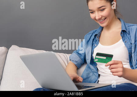 Online Shopping. Junge Frau sitzt auf einem Sofa studio isoliert auf Grau mit ec-Karte surfen Laptop lächelnd schließen aufgeregt - Bis Stockfoto