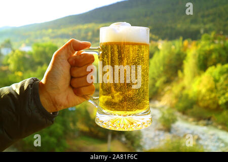 Des Menschen Hand mit einem Pint Bier gegen blurry Vorberg Waldblick in der Abendsonne Stockfoto