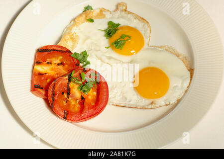 Morgen essen Spiegeleier aus zwei Eiern und Scheiben von gegrillten Tomaten auf eine weiße Platte, weißer Hintergrund Stockfoto