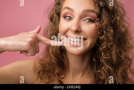 Closeup Schuß von hübsch Lächeln der kaukasischen Frauen Lächeln mit dem lockigen Haar das Tragen von Zahnspangen auf rosa Hintergrund isoliert. Stockfoto