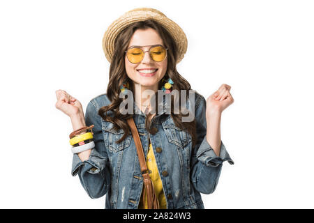 Aufgeregt boho Mädchen in der bootsfahrer und Sonnenbrille holding Fäuste oben mit geschlossenen Augen isoliert auf weißem Stockfoto