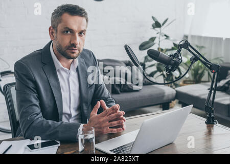 Stattliche radio Host auf Kamera beim Sitzen am Arbeitsplatz in der Nähe von Mikrofon und Laptop Stockfoto