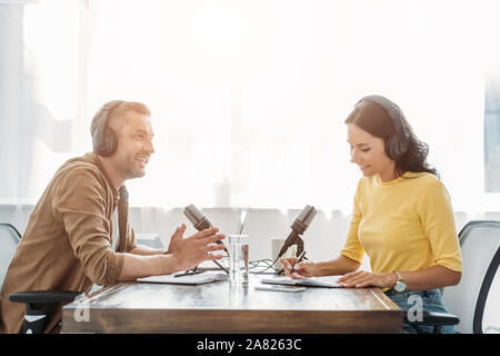 Zwei lächelnde radio Hosts in Kopfhörer sprechen beim Sitzen am Tisch im Studio Stockfoto