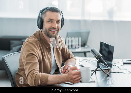 Stattliche radio host Lächeln auf die Kamera, während am Arbeitsplatz sitzen mit Tasse Kaffee Stockfoto