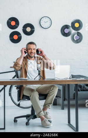 Stattliche radio host Lächeln auf die Kamera, während am Arbeitsplatz sitzen und das Setzen auf Kopfhörer Stockfoto