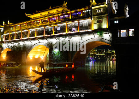 Die nächtliche Silhouette eines Mannes mit einem Sampan Boot schwimmt unter den Bögen der hell erleuchteten Phoenix Hong Brücke in Fenghuang antike Stadt in Stockfoto