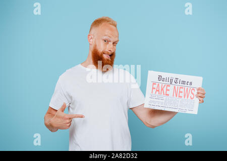 Lächelnd bärtiger Mann auf Zeitung mit gefälschten Nachrichten, auf Blau isoliert Stockfoto