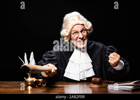 Wütend Richter in gerichtlichen Robe und Perücke am Tisch sitzen und halten bat isoliert auf Schwarz Stockfoto