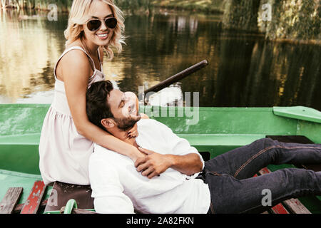 Attraktive Mädchen in Sonnenbrille umarmt glücklich Freund beim Sitzen im Boot auf dem See Stockfoto
