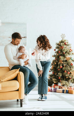 Mutter zeigt christmas Ball auf das Kind sitzt auf Väter Hände in der Nähe von geschmückten Weihnachtsbaum Stockfoto