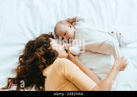 Blick von oben auf die Mutter berühren Bein von niedlichen kleinen Tochter beim Lügen auf weiße Bettwäsche Stockfoto