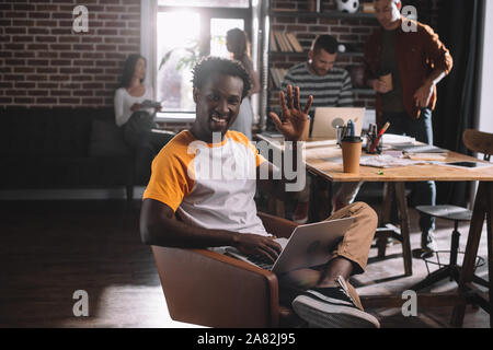 Lächelnd afrikanische amerikanische Geschäftsmann mit Laptop im Sessel sitzt und winkende Hand in der Nähe der jungen multikulturellen Kollegen Stockfoto
