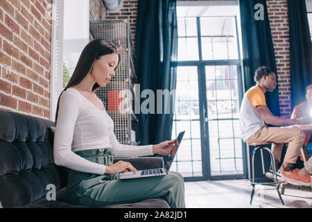 Nachdenklich Geschäftsfrau mit Laptop beim Sitzen auf einem Sofa in der Nähe von multikulturellen Kollegen Stockfoto