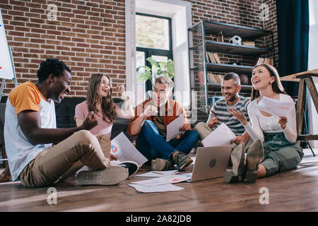 Jung, fröhlich multikulturellen Geschäftsleute sitzen auf dem Boden und zeigen Gewinner Gesten Stockfoto