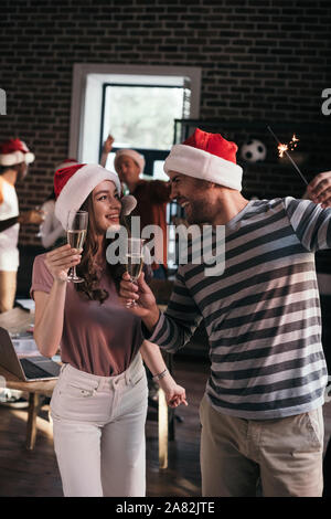 Junge, glücklich, Geschäftsmann und Geschäftsfrau in santa Hüte an einander suchen und dabei Champagner Gläser Stockfoto