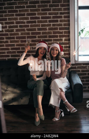 Zwei junge, glückliche Geschäftsfrau in santa Hüte Lächeln auf die Kamera beim Sitzen auf einem Sofa mit Wunderkerzen und Champagner Gläser Stockfoto