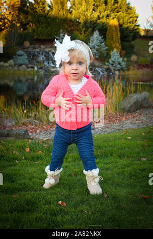 Sehr niedliches Kleinkind blonde Mädchen mit großen blauen Augen lächeln Mitten im schönen Herbst Park tragen Rosa Jacke und blaue Jeans und Stockfoto