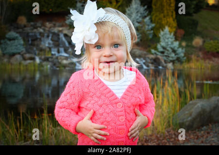 Sehr niedliches Kleinkind blonde Mädchen mit großen blauen Augen lächeln Mitten im schönen Herbst Park tragen Rosa Jacke und blaue Jeans und Stockfoto