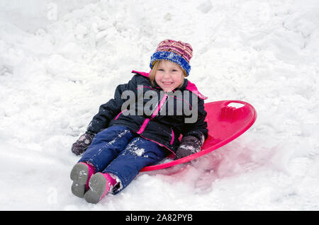 Ein süßes kleines Mädchen sitzt auf ihrem roten Untertasse Schlitten im Schnee. Was für eine tolle Möglichkeit, den Winter zu genießen! Stockfoto