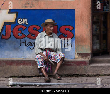 Eine ältere Maya Mann in die traditionelle Kleidung der San Pedro La Laguna, Guatemala, sitzt mit einer gepflasterten Straße vor einem Geschäft. Stockfoto