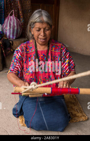 Eine ältere, grauhaarige Frau Maya webt Stoff auf einem Backstrap Loom kniend auf dem Boden ihres Hauses in Santa Catarina Palopo, Guatemala. Stockfoto