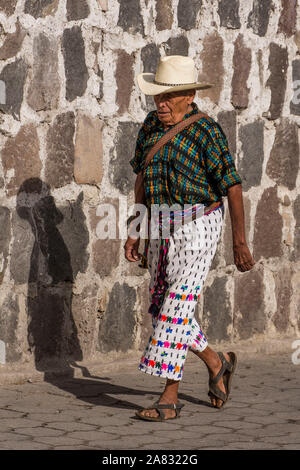 Eine ältere Maya Mann in traditioneller Kleidung von San Pedro La Laguna, Guatemala, in der Straße mit Kopfsteinpflaster vor einer groben Stein Wand geht. Stockfoto
