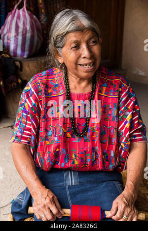 Eine ältere, grauhaarige Frau Maya webt Stoff auf einem Backstrap Loom kniend auf dem Boden ihres Hauses in Santa Catarina Palopo, Guatemala. Stockfoto