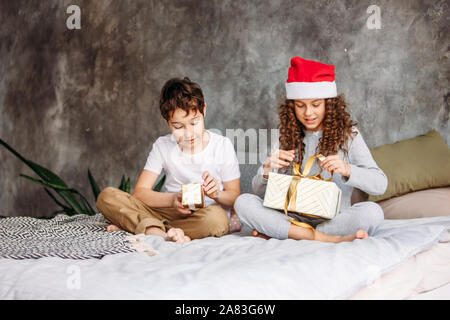 Cute tween Kinder in Santa Hüte und Schlafanzüge geöffnet Weihnachten Geschenkboxen auf dem Bett mit Kissen, Weihnachten Morgen Zeit, Kinder Partei Stockfoto