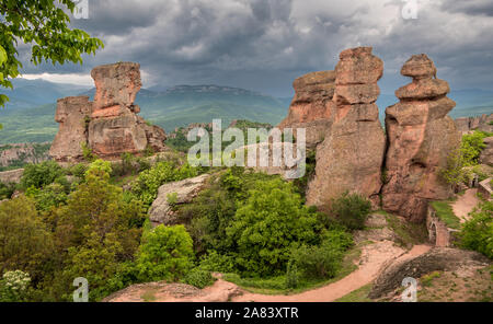 Schöne Landschaft mit bizarren Felsformationen, üppig grüne Vegetation, Himmel mit Gewitterwolken und Bergen im Hintergrund, belogradchik Felsen. Stockfoto