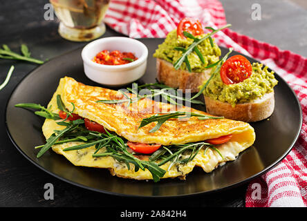 Frühstück. Omelett mit Tomaten, Käse, grün Rucola und Toast mit Avocado Creme auf schwarze Platte. Frittata - italienische Omelette. Stockfoto