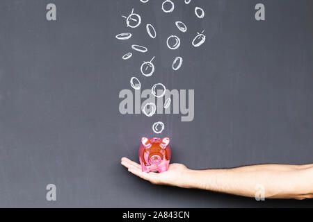 Konzept der Geld sparen mit einem rosa Sparschwein in der Hand vor einer Tafel mit Hand gezeichnet Münzen statt Stockfoto