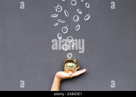 Konzept der Geld sparen mit einem goldfarbenen Sparschwein in der Hand vor einer Tafel mit Hand gezeichnet Münzen statt Stockfoto