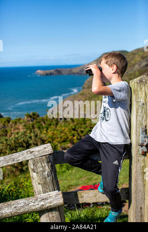 Ein kleiner Junge, der auf einem Holzzaun sitzt, blickt durch sein Fernglas aufs Meer, Wales, Großbritannien. Stockfoto