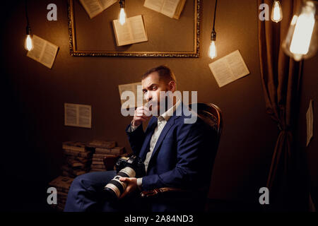 Portrait von Stattlichen bärtiger junger Mann mit blauen Anzug, weißes Hemd in Luxus Bibliothek Halten der Kamera sitzen. Fotograf, Videofilmer. Stockfoto