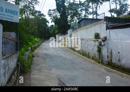 Die geschwungene Straße entlang einer hohen Mauer in den Hügeln von Shillong mit vielen Bäumen und Grünflächen Stockfoto