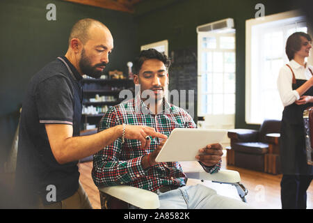Männliche Friseur- und Kunden mit digitalen Tablet sprechen in Barbershop Stockfoto