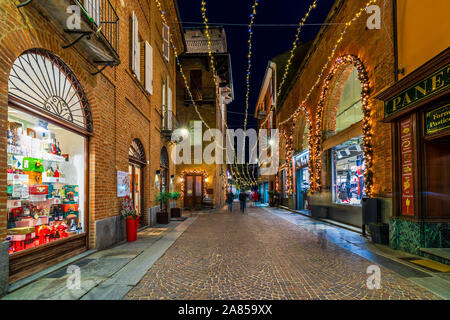 Schmale Straße mit Kopfsteinpflaster und Geschäfte mit Weihnachtsbeleuchtung in Abend in Alba, Italien eingerichtet. Stockfoto