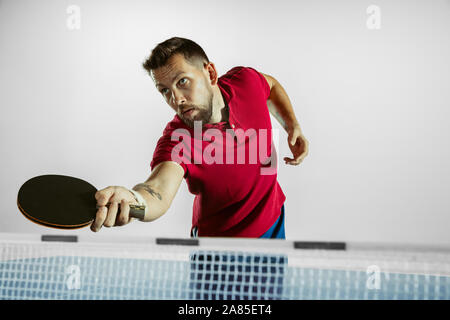 Stark. Junger Mann spielt Tischtennis auf weißem Studiohintergrund. Modell spielt Ping pong. Konzept der Freizeitgestaltung, Sport, menschliche Emotionen in Gameplay, gesunden Lebensstil, Bewegung, Aktion, Bewegung. Stockfoto