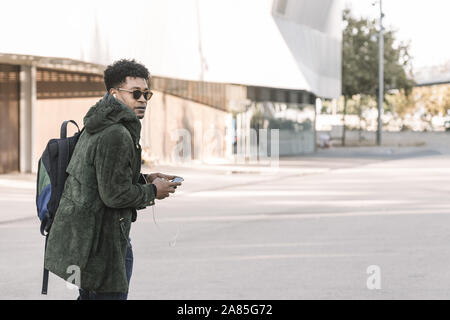Afrikanische amerikanische junge männliche mit Kopfhörern zu Fuß durch die Stadt und hören Musik mit einem Mobiltelefon, Technologie und lifestyle Konzept über interne Stockfoto