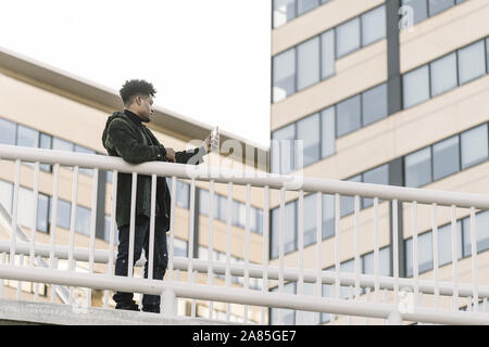 Schweren afrikanischen schwarzen Mann mit Ohrhörer, Videokonferenz mit einem Mobiltelefon lehnte sich auf einem Geländer im Freien in der Stadt, Technologie und Lifestyle co Stockfoto