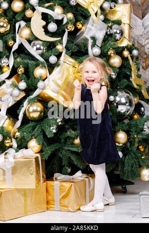 Portrait von freudiger elementare Alter Mädchen mit blauen Kleid stanging neben Weihnachtsbaum glücklich zu Weihnachtsgeschenke erhalten Stockfoto