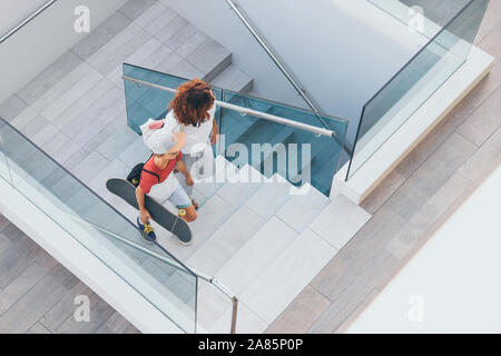 Trendy Mutter und Sohn zu Fuß auf der Treppe in einer modernen Stadt. Junge mit Skateboard geht auf die Leiter mit Mama. Blick von oben auf die Menschen Spaziergänge Stockfoto