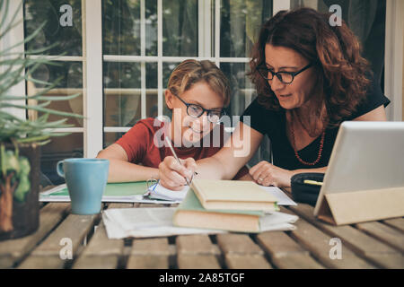 Junge Schüler Hausaufgaben zu Hause mit Schule Bücher, Zeitung und digitale Pad von seiner Mutter geholfen. Mom schreiben auf dem copybook Lehre seinen Sohn. Stockfoto