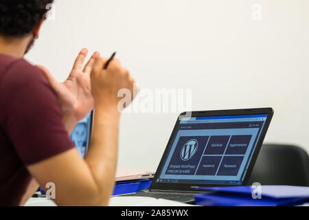 Laptop mit dem Wordpress Logo auf dem Bildschirm fotografiert, junge Fahrer teilweise von hinten mit einem Stift in der Hand gerahmt. Stockfoto