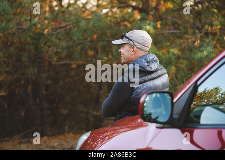 Ein Mann lehnt sich auf der Motorhaube eines Autos. Der Blick von der Rückseite. SUV im Wald. Mann mit verschränkten Armen lehnte sich zurück auf Auto. Stockfoto