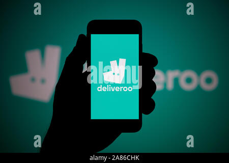 Ein Mann schaut auf seinem iPhone die zeigt die Deliveroo Logo, mit dem gleichen Logo im Hintergrund (nur redaktionelle Nutzung). Stockfoto