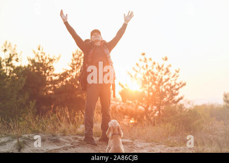 Glücklich müde Reife touristische heben die Hände hoch. Älterer Mann mit Rucksack gegen Abend Sonne und seinem Cocker Spaniel sitzen in der Nähe von Sand. Stockfoto