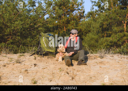 Tourist mit Rucksack und Hund haben Pause im Wald. Ein Mann lächelt, hält einen Hund Pfote in seinen Händen. Stockfoto