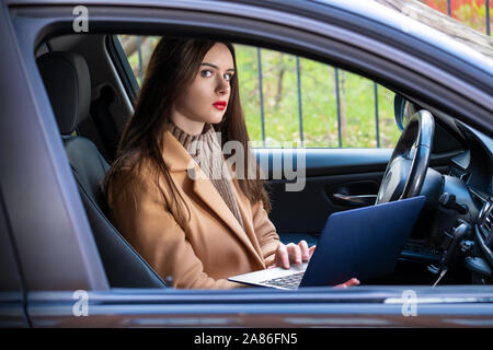 Junge Frau sitzt in ihrem Auto mit einem Laptop in den Händen. Sie ist ernst und auf der Suche nach etwas im Internet. Konzept der Autoversicherung und roadsid Stockfoto