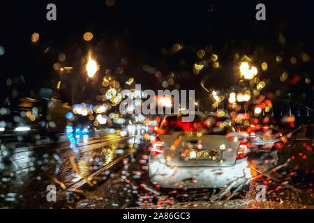 Bild des Autoverkehrs im Regen in den späten Abend, Schuß durch ein Glas mit Regentropfen. Abstraktes Bild mit Farbflecken. Stockfoto
